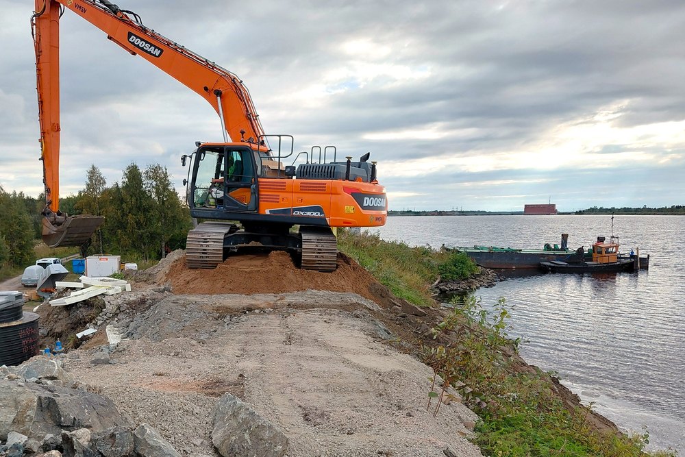 Pitkäpuominen kaivinkone joen penkereellä ja hinaaja vesillä